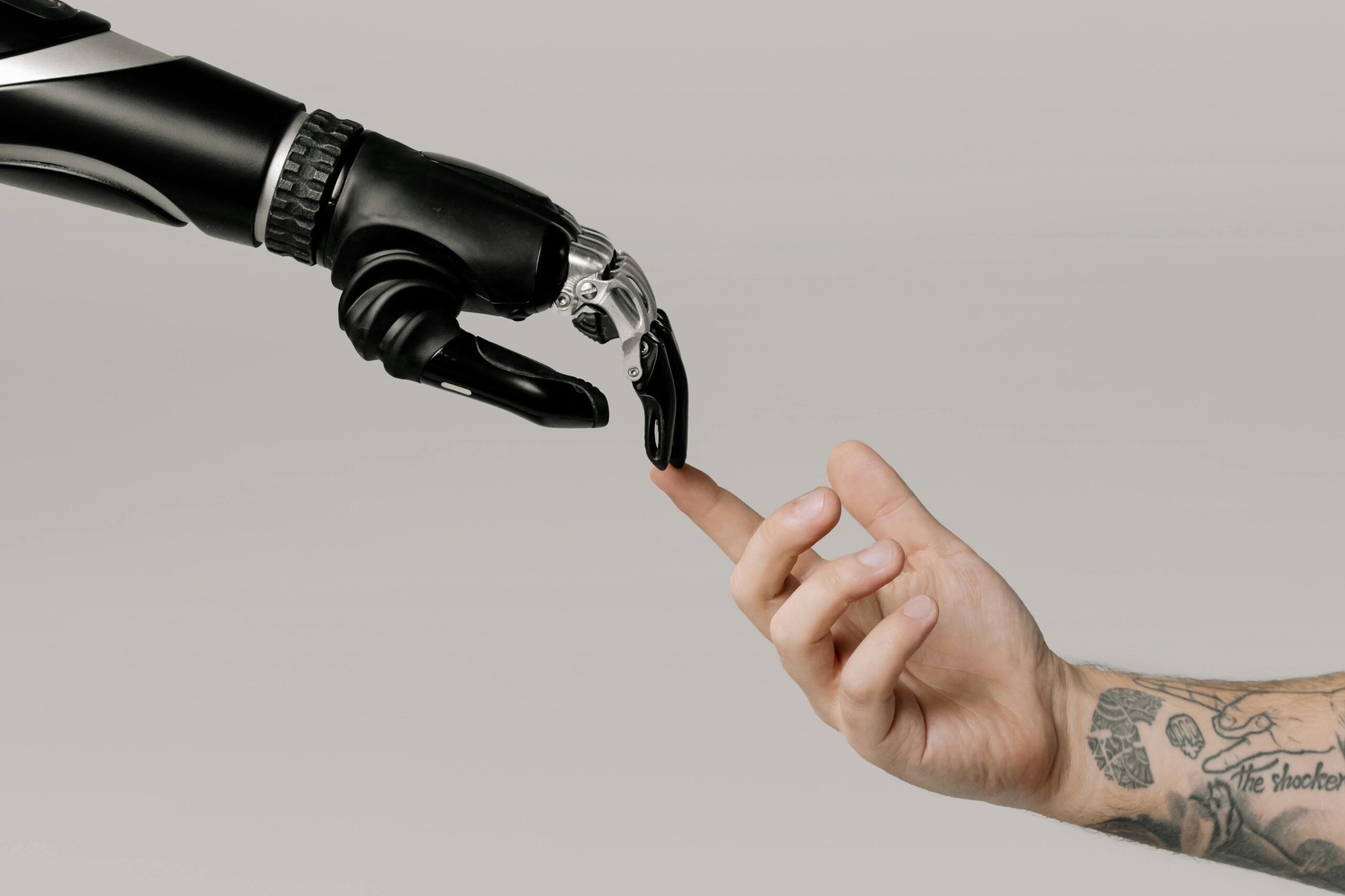 Home A human hand with tattoos reaching out to a robotic hand on a white background.